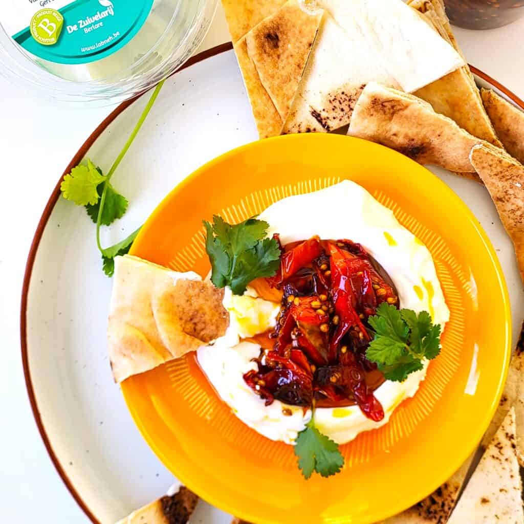 Labneh met gekarameliseerde paprika's, verse kruiden en pita op een gele schaal.